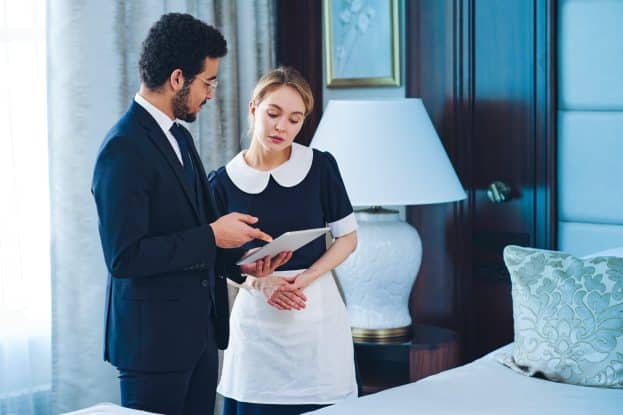 Confident owner of luxurious hotel giving instructions to chambermaid while pointing at tablet screen with official duties of service staff