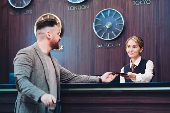 Young female receptionist standing by counter in lounge and looking at male client while passing him passport after checking it in